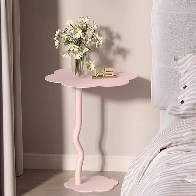 a small pink table with flowers in a vase on it next to a white bed