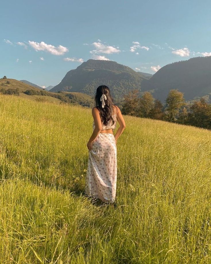 a woman standing in the middle of a field with her hands on her hips looking at mountains