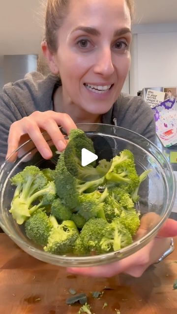 a woman holding a bowl full of broccoli on top of a wooden table