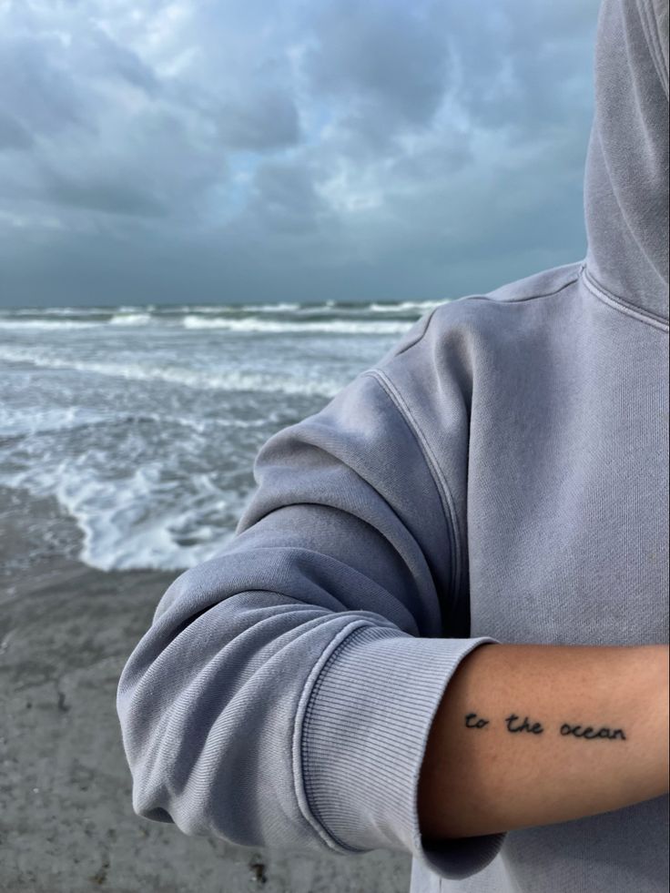 a person with a tattoo on their arm standing in front of the ocean and cloudy sky