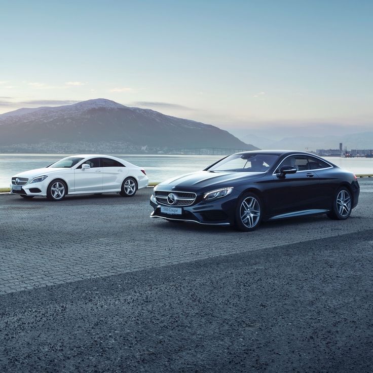 two mercedes cars parked next to each other on the road near water with mountains in the background