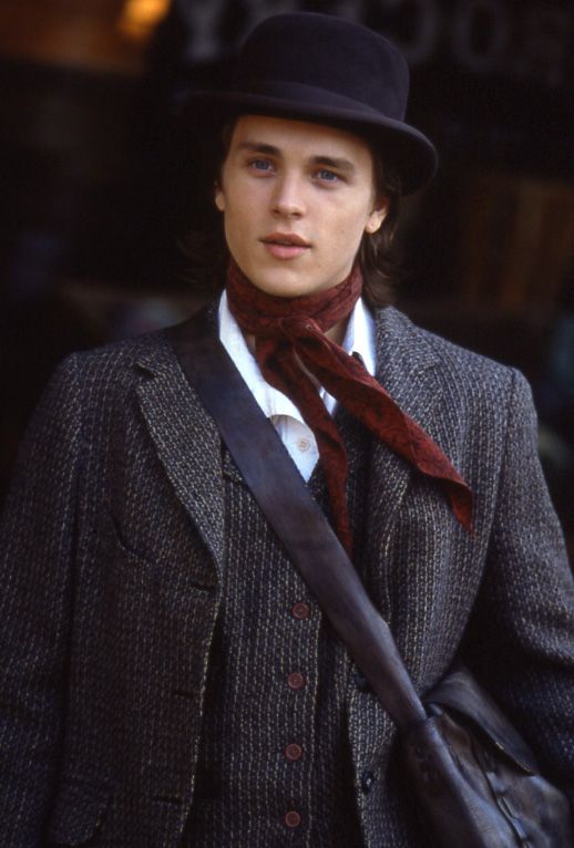 a man wearing a suit and tie with a hat on his head is standing in front of a building