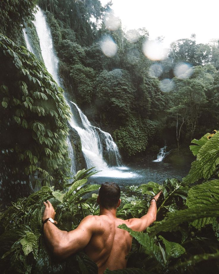 a man standing in front of a waterfall surrounded by lush green plants and trees with his back turned to the camera