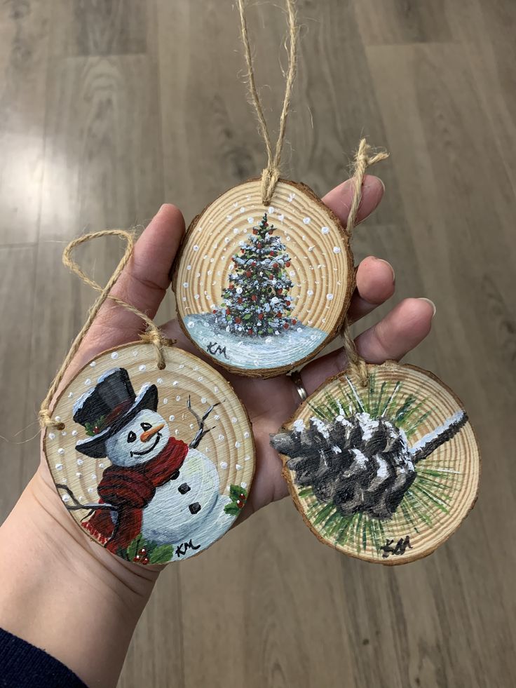 hand holding three christmas ornaments in front of a wood flooring background with a tree and snowman