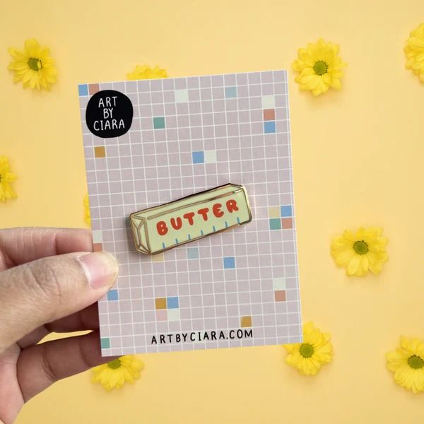 a hand holding a pin with the word butter on it in front of yellow flowers