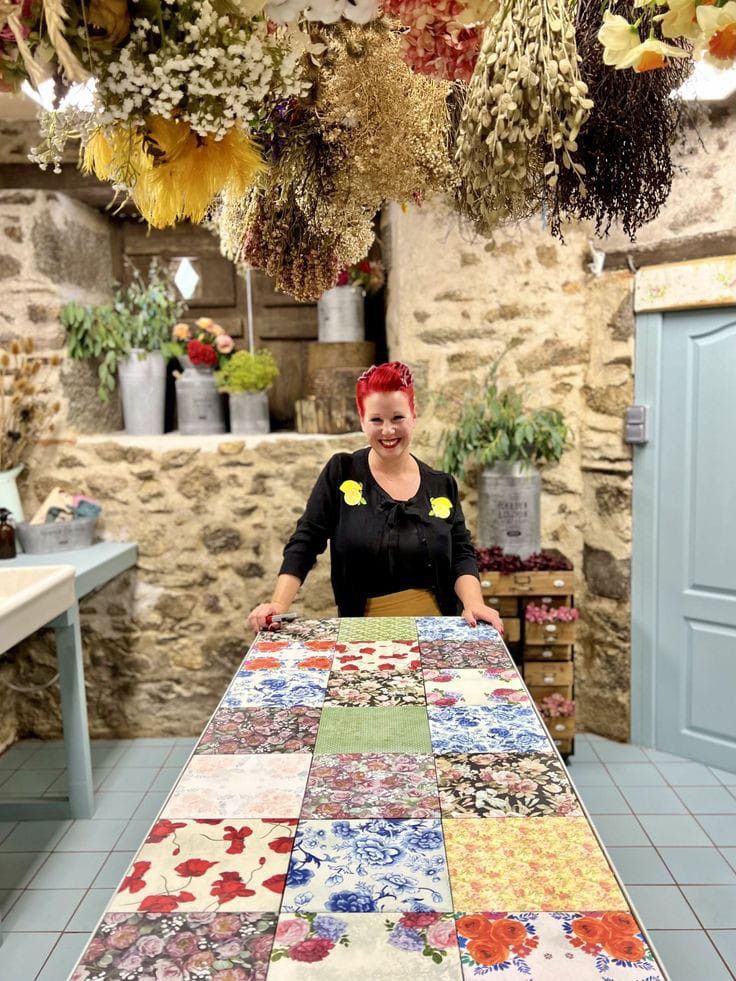 a woman standing behind a table covered in lots of different types of fabric and flowers