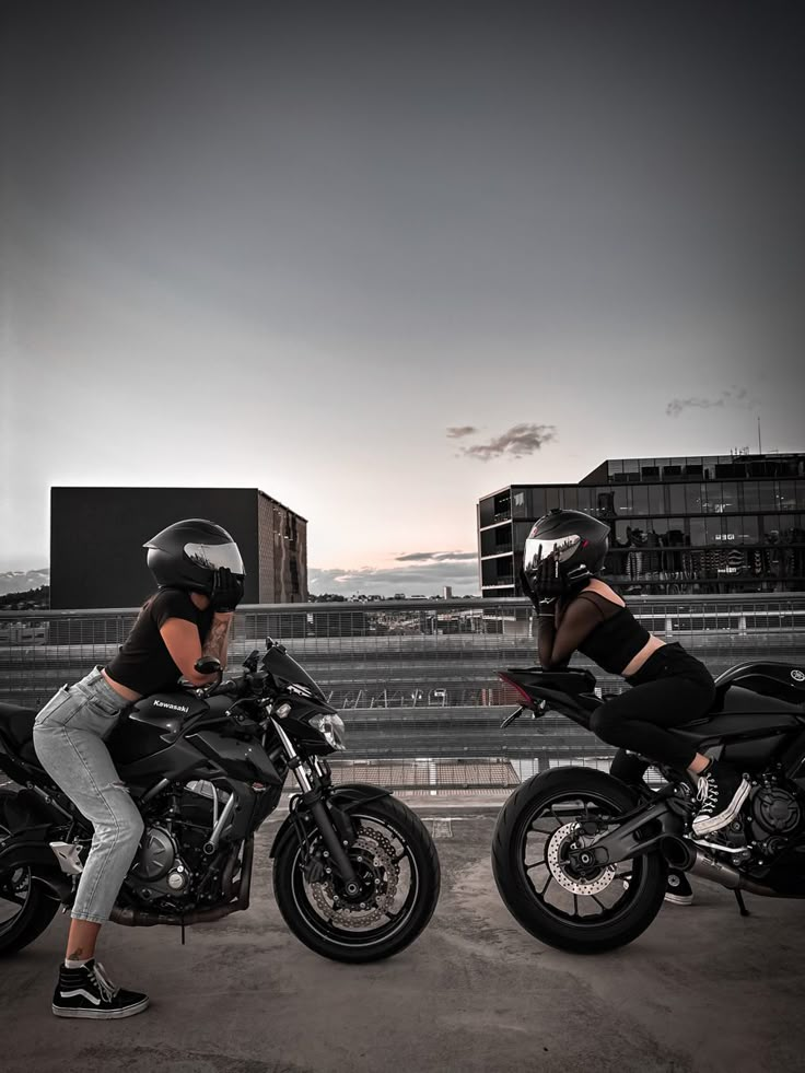 two women sitting on motorcycles in front of a cityscape with buildings behind them