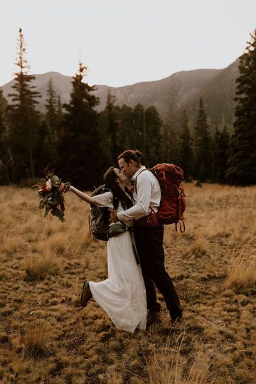 a bride and groom are walking through the field with backpacks on their backs, kissing