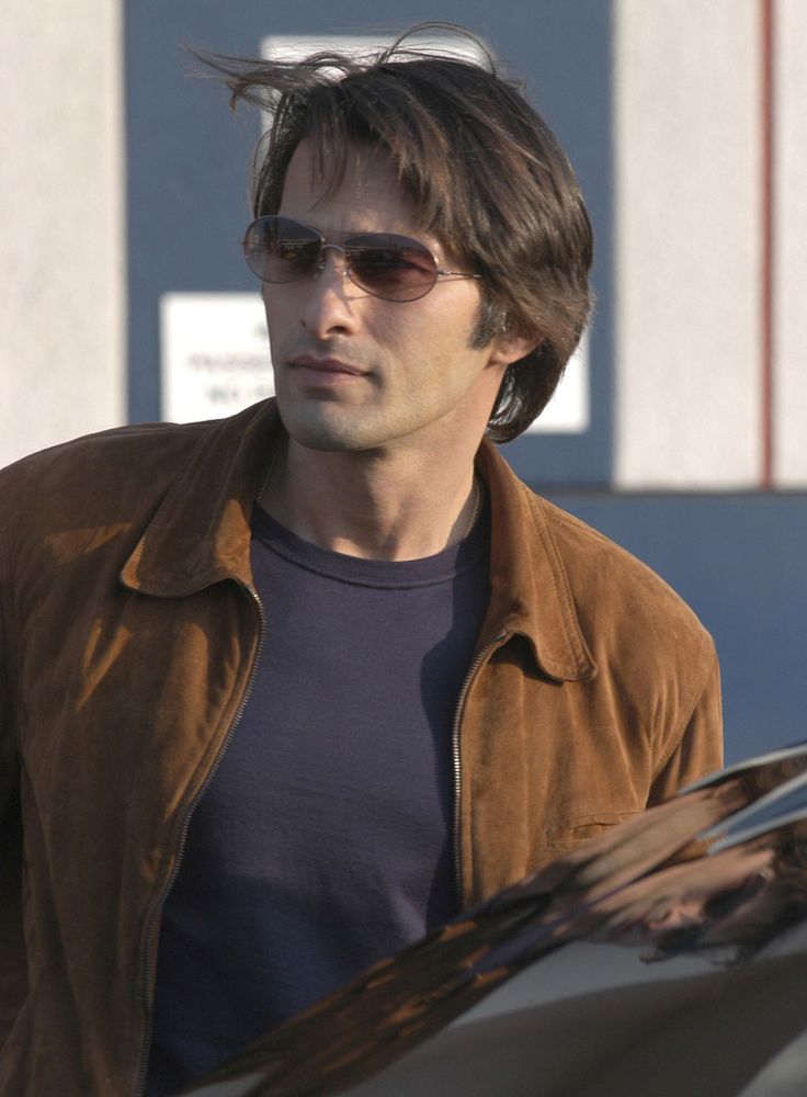 a man with sunglasses and a brown jacket is standing in front of a parked car