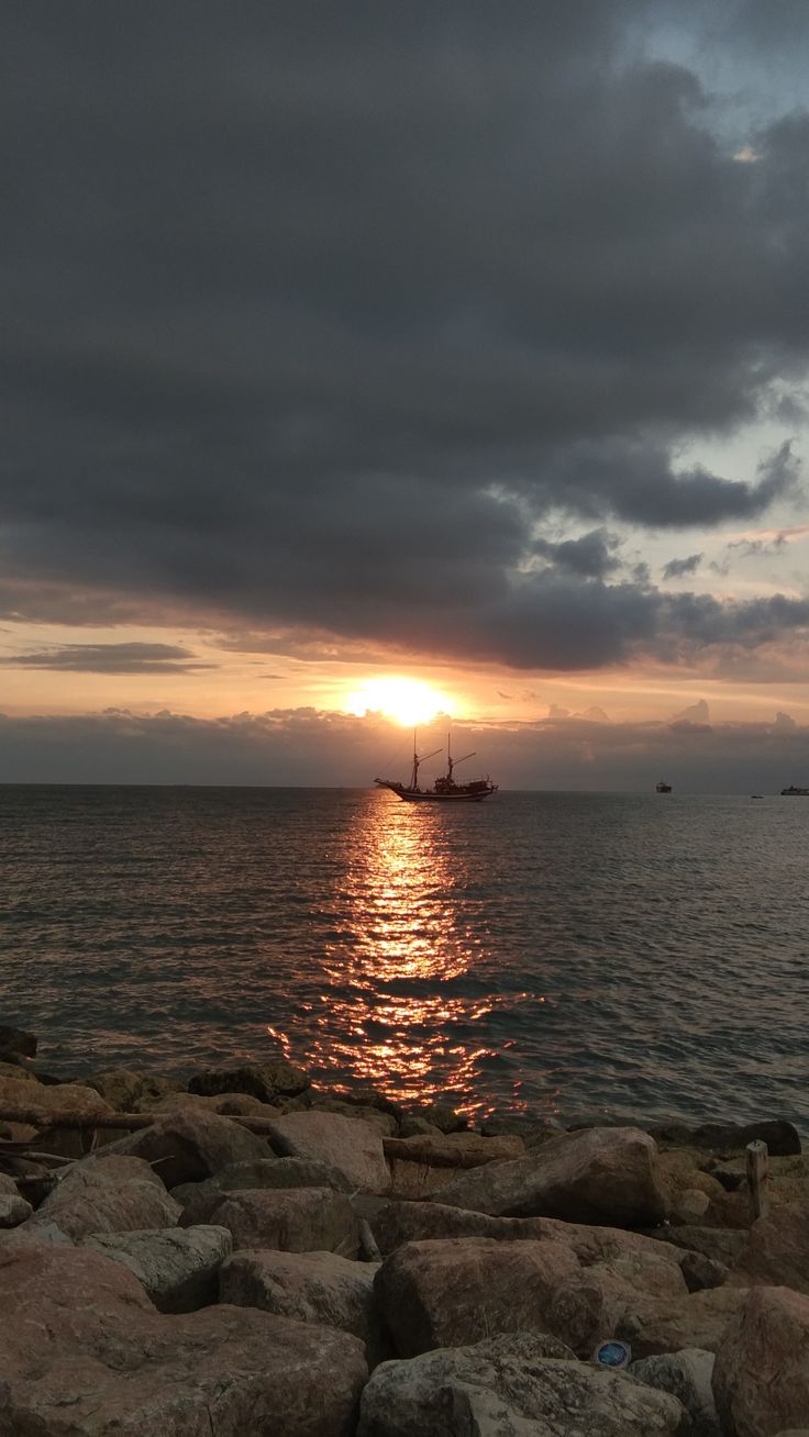 the sun is setting over the ocean with rocks on the shore and boats in the water