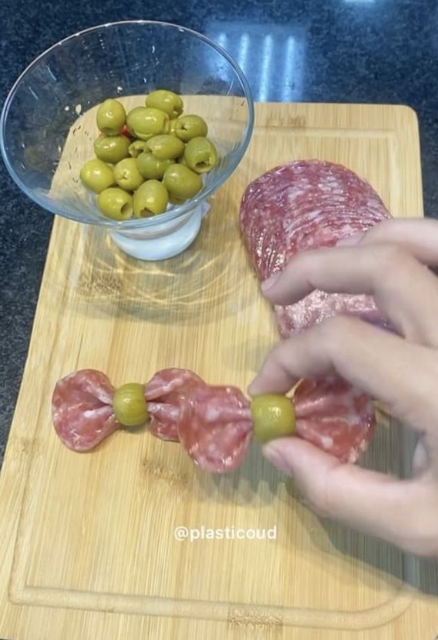 someone is cutting up some meat and olives on a wooden board with a bowl of green olives