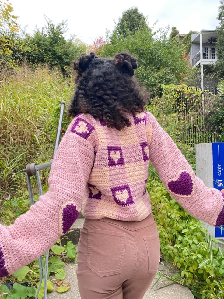 a woman wearing a pink and purple knitted sweater with hearts on the sleeves, standing in front of a garden