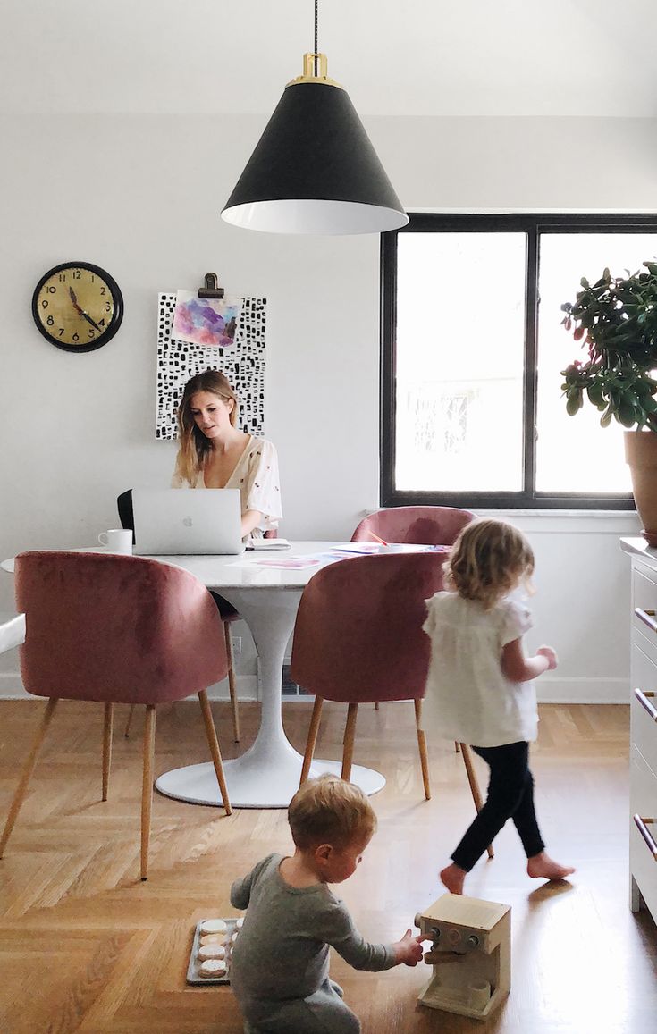 two small children playing in the middle of a room with a table and chairs, while a woman works on her laptop