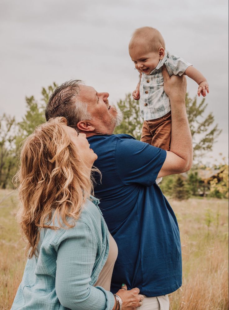 a man holding a baby up to his face while standing next to a woman in a field