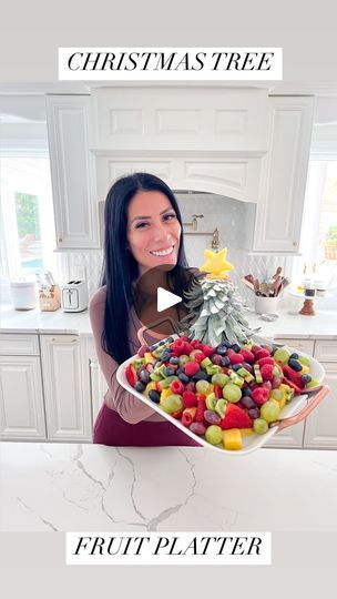 a woman holding a platter full of fruit in front of a christmas tree on top of a kitchen counter