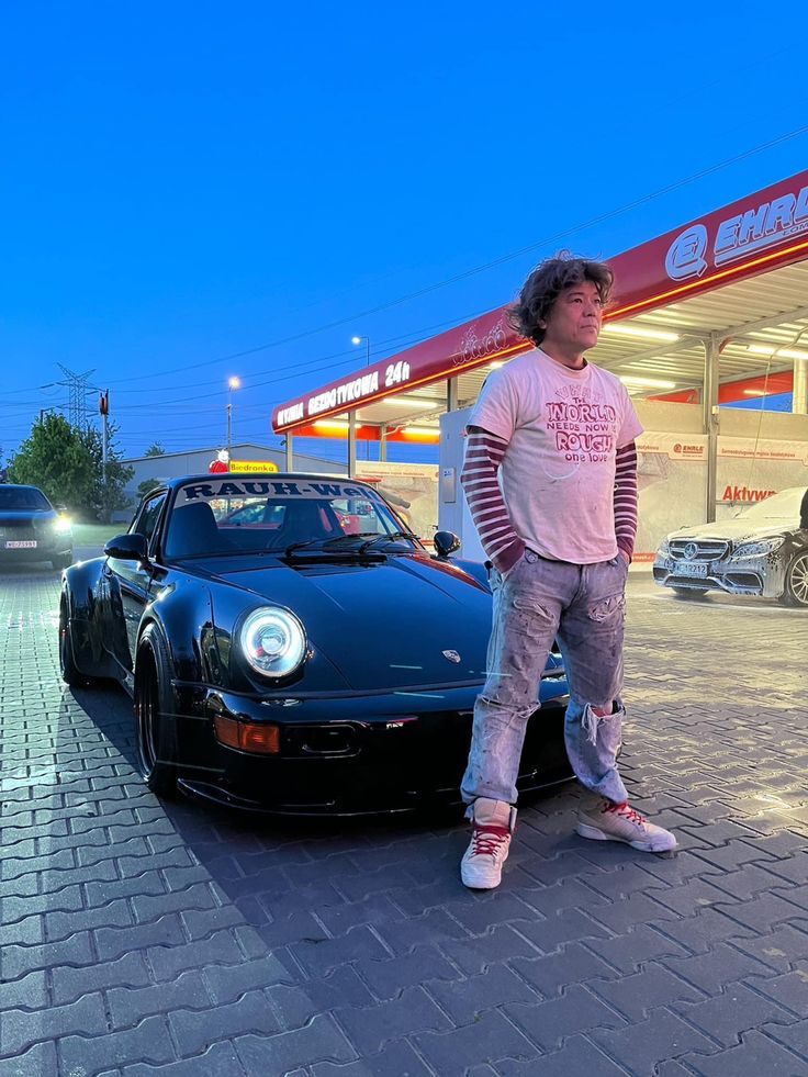 a man standing next to a black sports car in front of a gas station at night