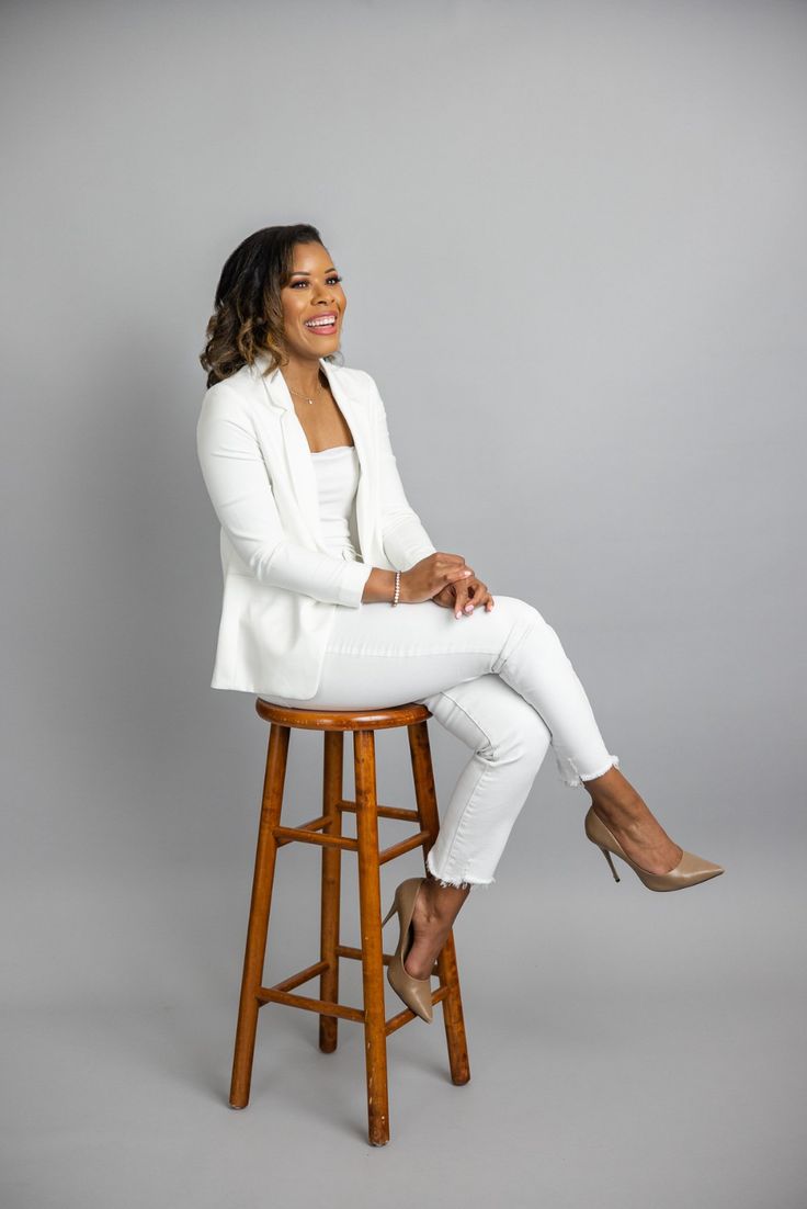 a woman sitting on top of a wooden stool wearing white pants and blazer jacket