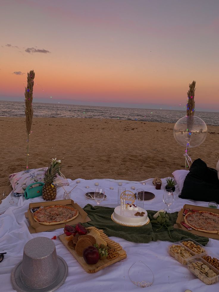 a table with food and drinks on it at the beach near the ocean during sunset