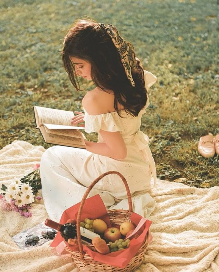 a woman sitting on a blanket reading a book next to a basket full of fruit