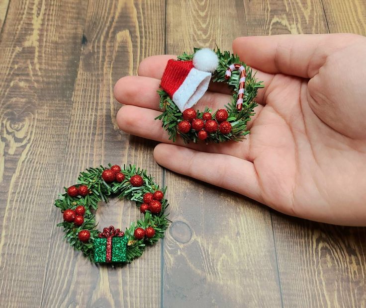 a hand holding a small christmas wreath on top of a wooden table next to a fake santa hat