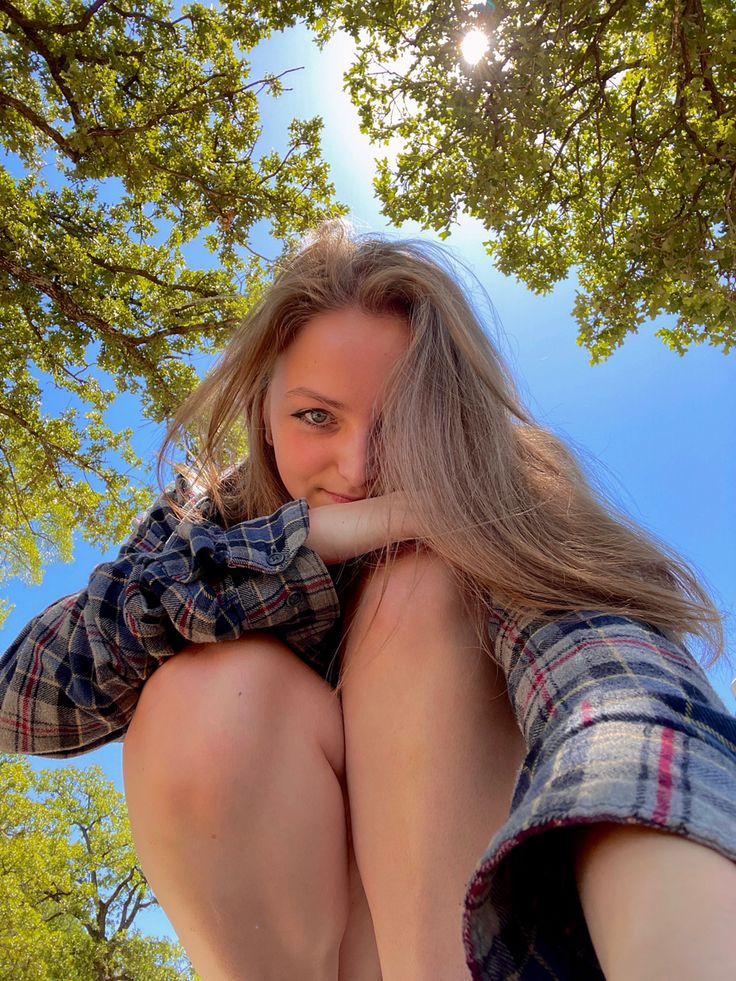 a woman sitting under a tree with her hands on her knees and looking at the camera