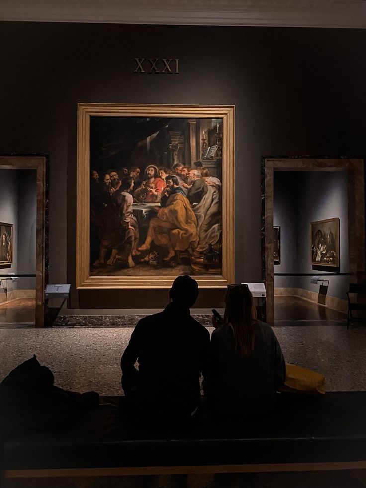 two people are sitting on a bench looking at paintings in an art gallery, with one person taking a photo