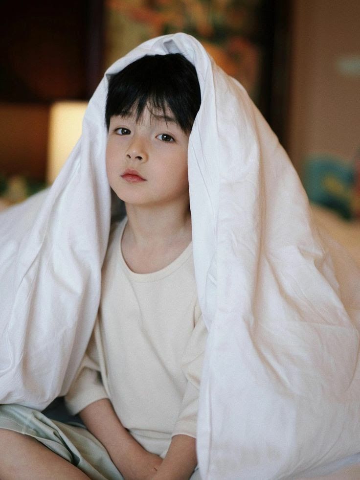 a young boy sitting under a white blanket on top of a bed in a bedroom