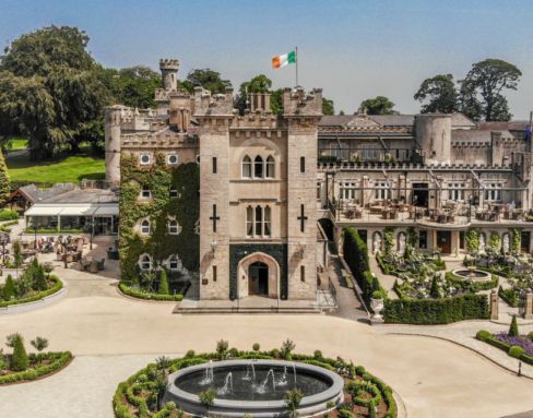 an aerial view of a castle like building surrounded by greenery and trees, with a fountain in the foreground