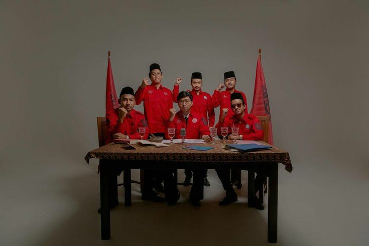 a group of men in red shirts sitting at a table with flags on the wall behind them