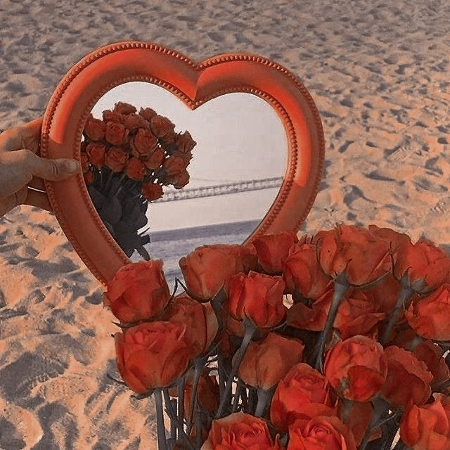 a heart shaped mirror sitting on top of a sandy beach next to flowers and a vase filled with red roses