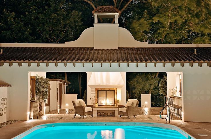 an outdoor swimming pool at night with lights on the ceiling and chairs around it in front of a house