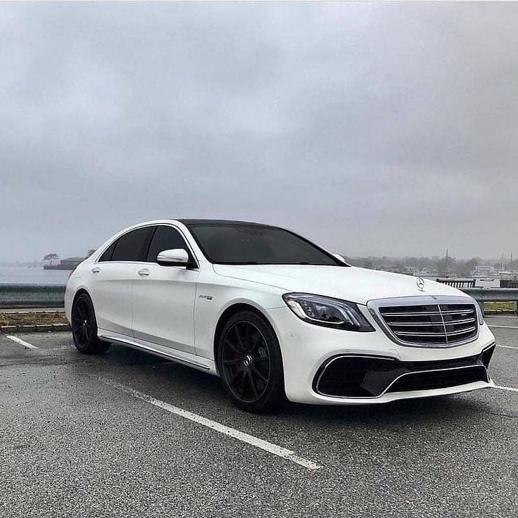 a white mercedes s - class parked in a parking lot next to the ocean on a cloudy day