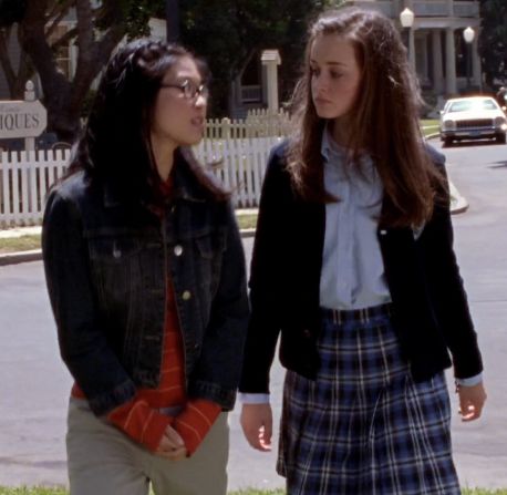 two young women standing next to each other on a sidewalk in front of a white picket fence