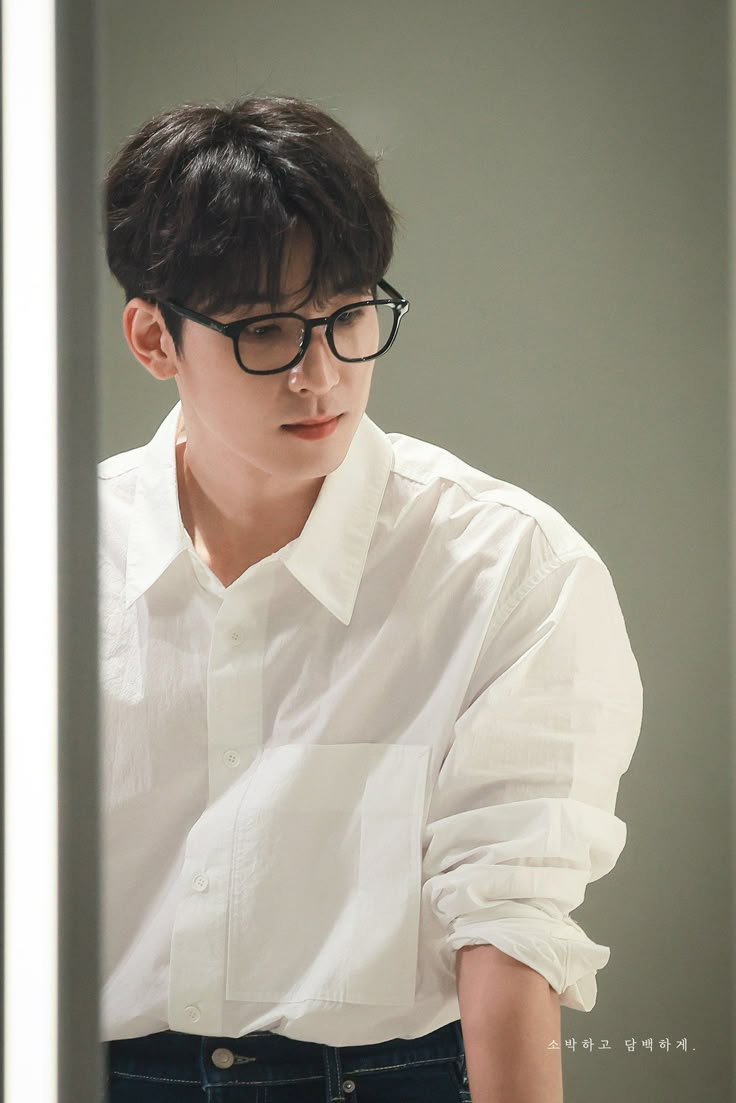 a young man wearing glasses and a white shirt looking at his cell phone while standing in front of a mirror