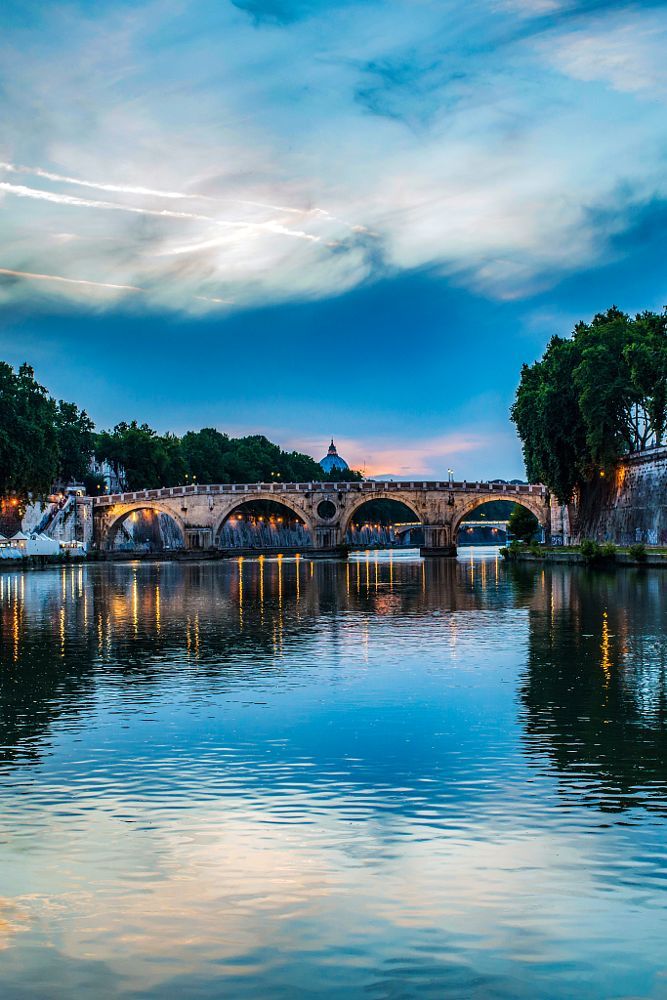 a bridge over a body of water with trees on both sides and clouds in the sky