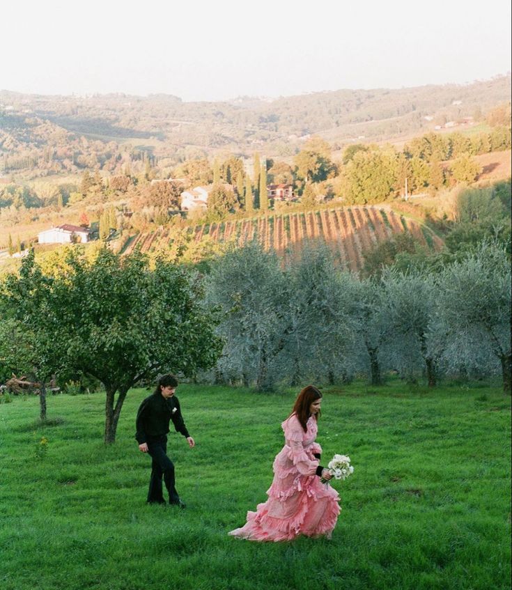 a man and woman are walking through the grass in an apple orchard with trees behind them