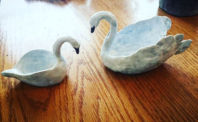 two white ceramic swans sitting on top of a wooden table next to eachother