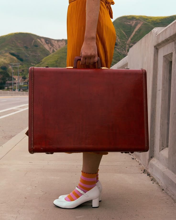 a woman in yellow dress holding a brown suitcase on sidewalk next to wall and mountains