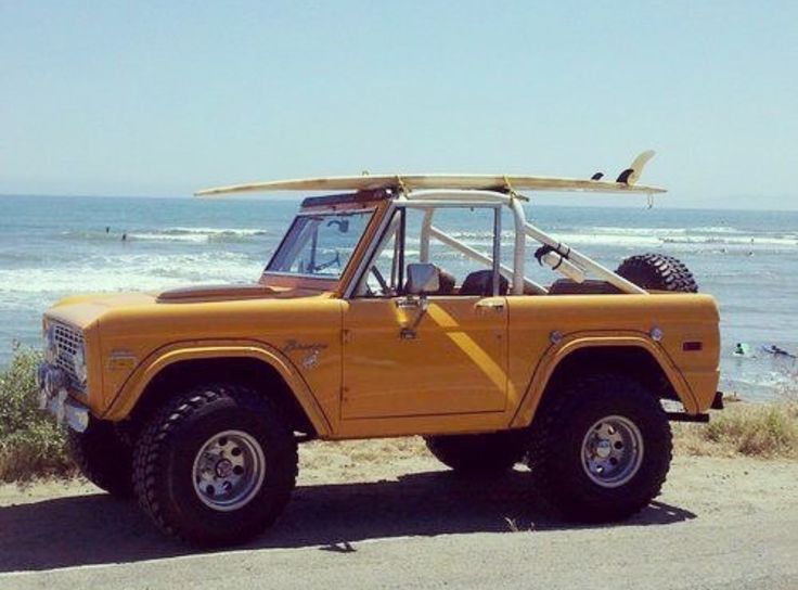 an old yellow jeep with a surfboard on top parked in front of the ocean