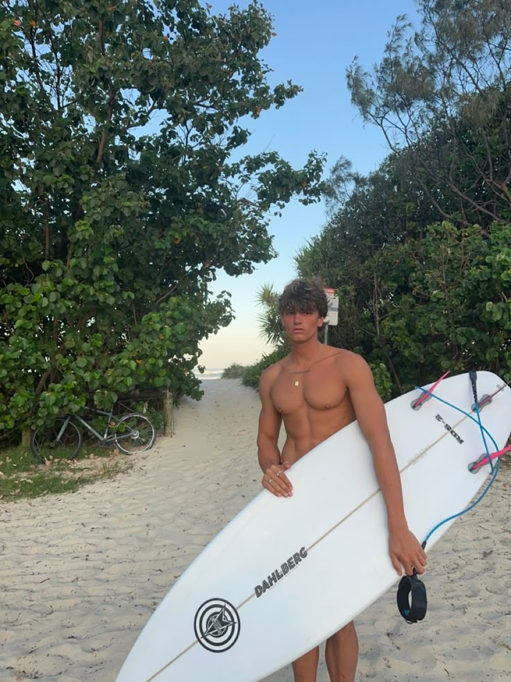 a shirtless man holding a white surfboard on the beach with trees in the background