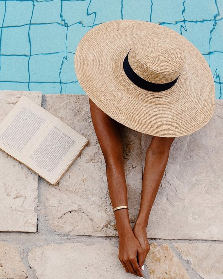 a woman sitting on the ground next to a pool wearing a hat and reading a book