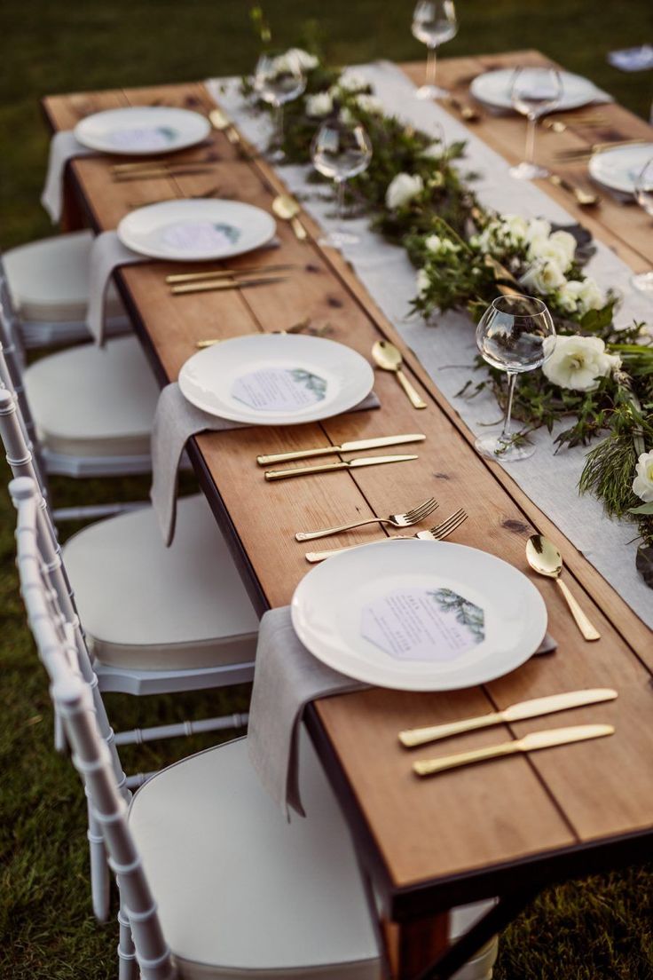 a long table set with white plates and silverware for an outdoor wedding reception in the grass