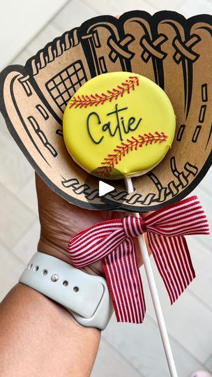 a person is holding a baseball themed cookie on a stick with the word cutie in it