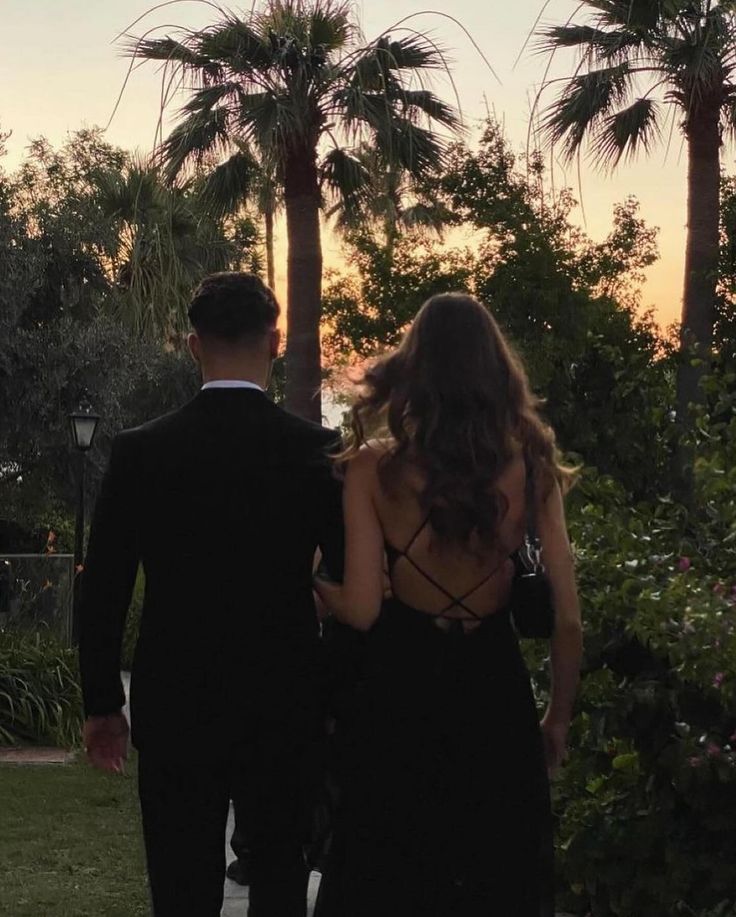 a man and woman in formal wear walking down a sidewalk at sunset with palm trees behind them