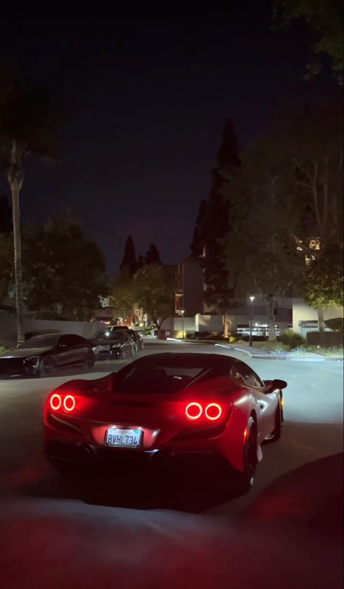 a red sports car is parked in the parking lot at night with its lights on