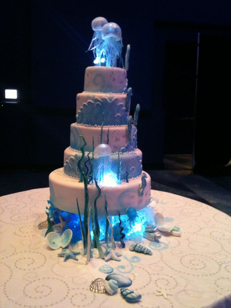 a three tiered wedding cake with blue lights on the side and sea shells around it