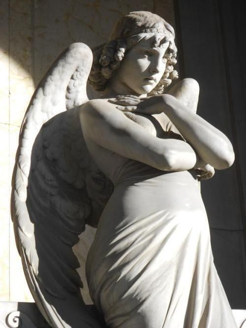 an angel statue sitting on top of a white wall next to a building with its arms crossed