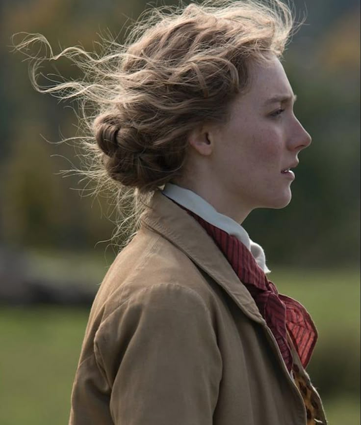 a woman with long hair wearing a brown jacket and red scarf standing in front of a green field