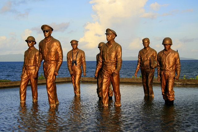 four bronze statues of men standing in water near the ocean with blue sky and clouds behind them