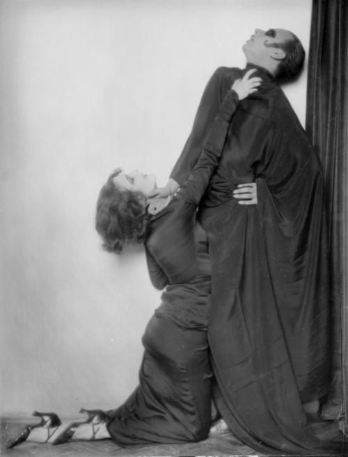 an old black and white photo of two people in dress clothes leaning against a curtain